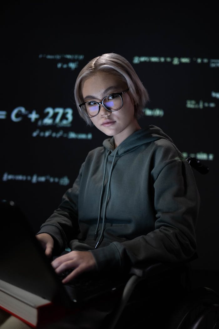 Young woman in glasses coding on laptop with projected math equations in a dimly lit room.